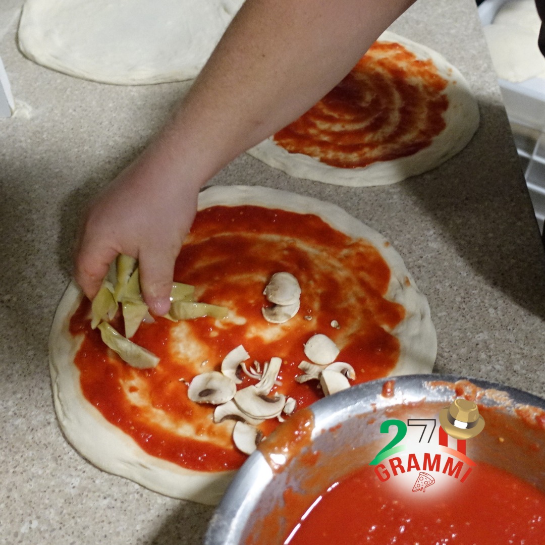 Mano che distribuisce carciofini e funghi champignon su pizze stese col pomodoro, logo 270 Grammi sulla ciotola di salsa