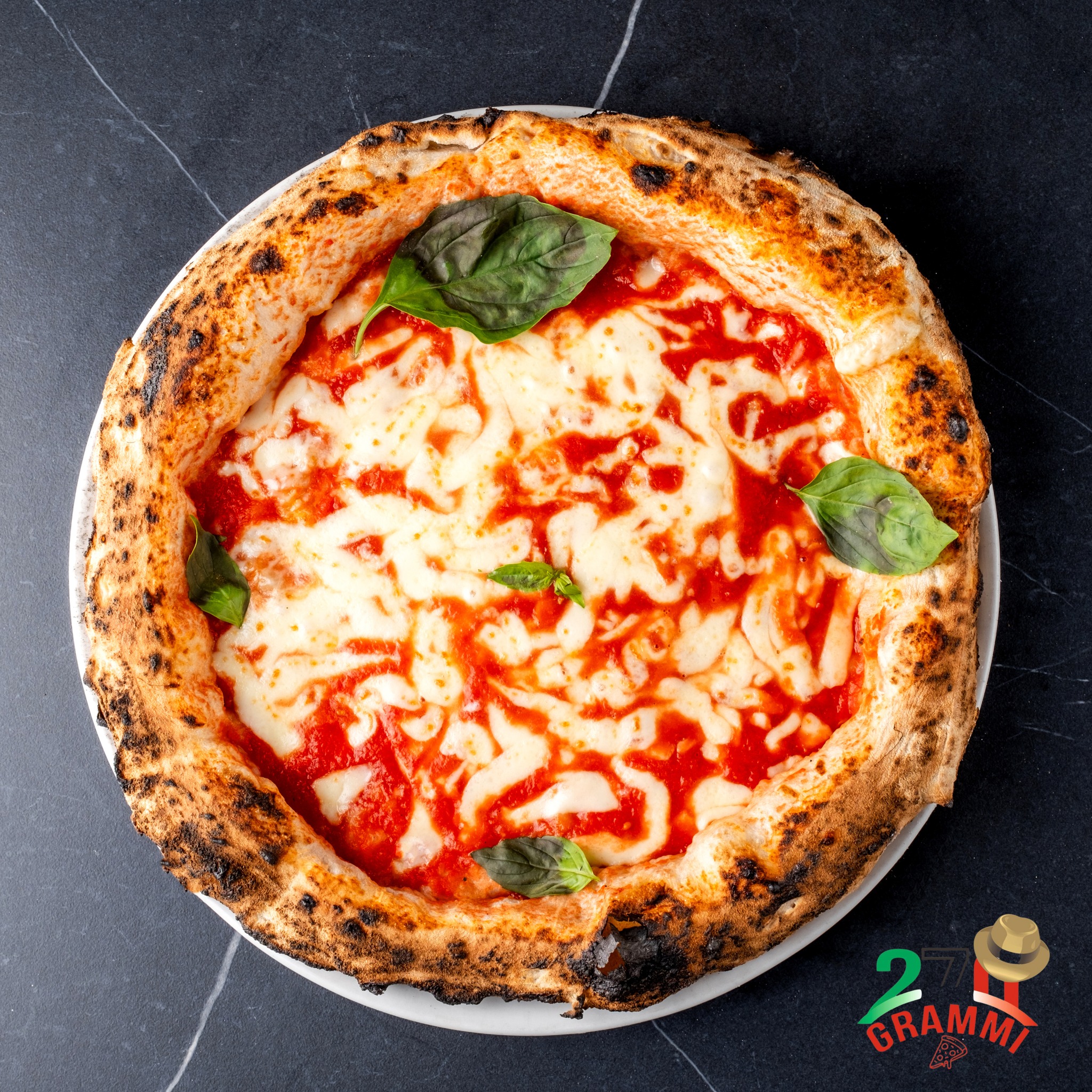Pizza margherita di 270 Grammi con cornicione alto bruciacchiato, mozzarella fusa e basilico su piatto bianco e marmo nero